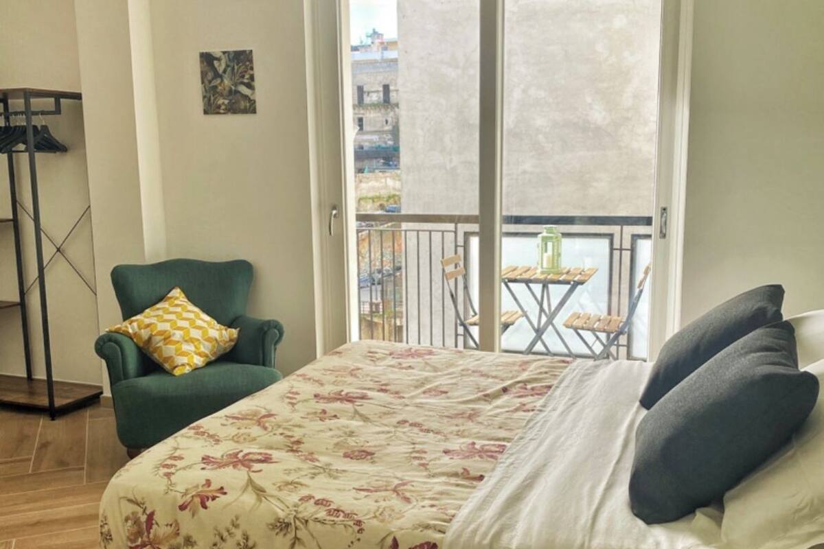 Un dormitorio con cama, una silla, un balcón con mesa y sillas, y vistas a edificios.