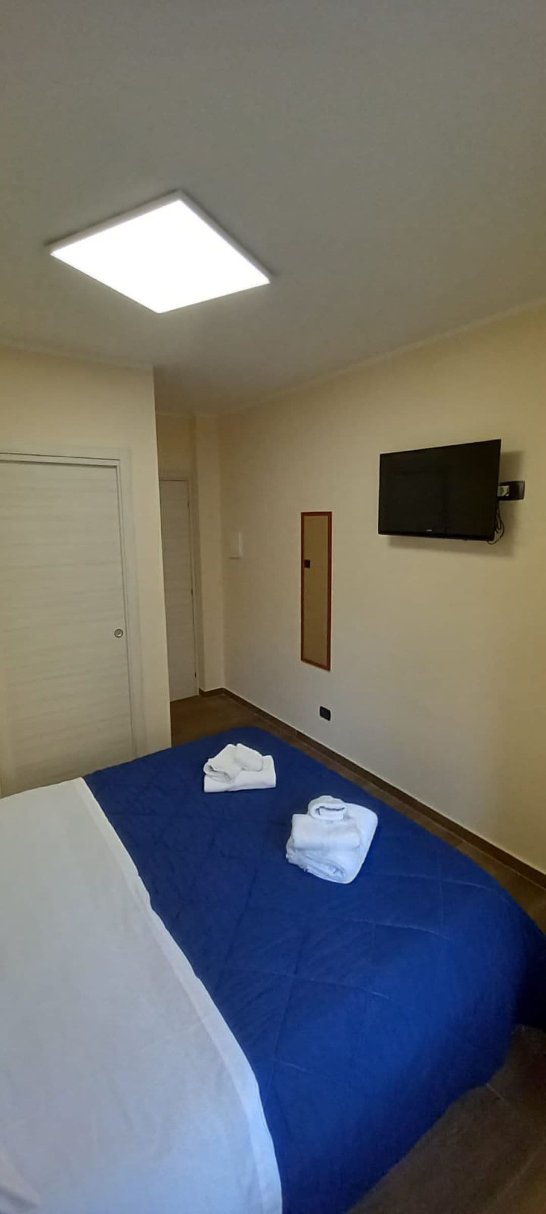 A hotel room with a bed, a flat-screen TV mounted on the wall, and a door.