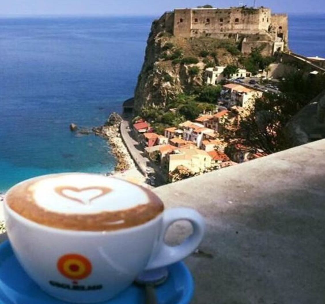 A cup of coffee with a heart design on a saucer, overlooking a coastal town with a historic castle on a cliff.