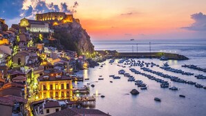 A coastal town at sunset with historic buildings, a fortified structure on a cliff, and a marina with numerous boats. - B&B Omnia Scilla (Scilla)
