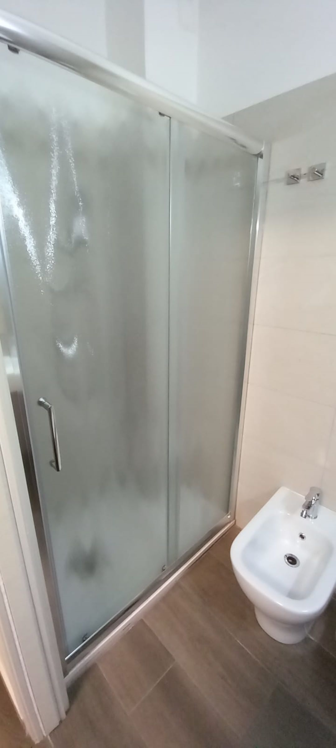 A bathroom with a glass shower enclosure, a white bidet, and tiled flooring.