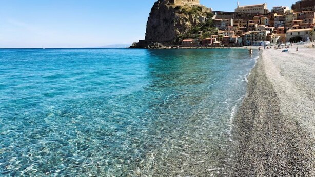 A coastal town with a prominent castle on a cliff, clear turquoise waters, and a pebble beach. - B&B Omnia Scilla (Scilla)
