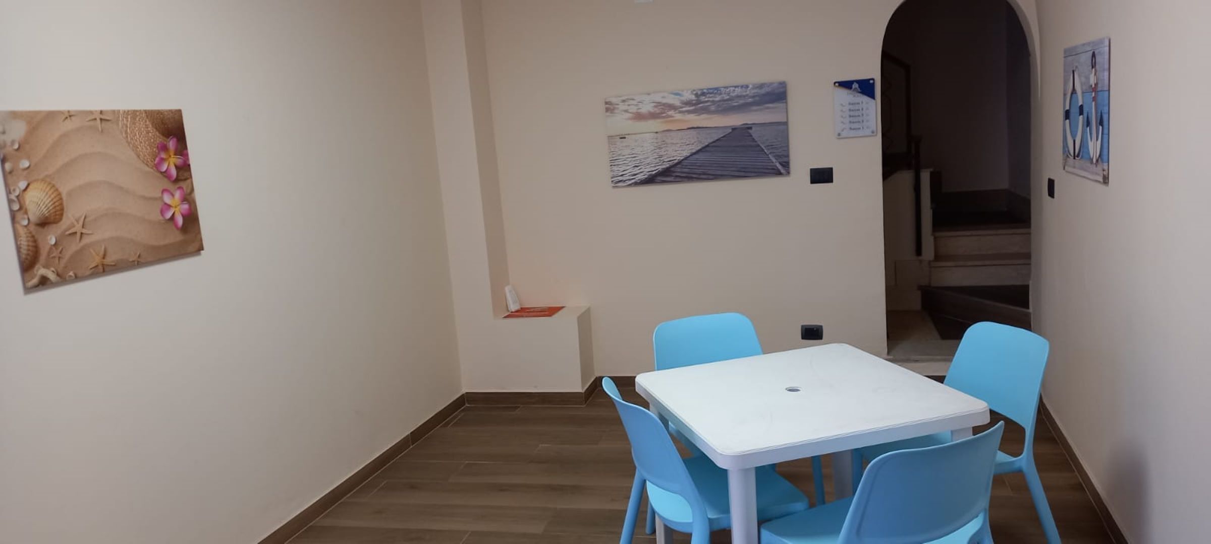 Una stanza modesta con un tavolo bianco e quattro sedie blu, un quadro raffigurante una spiaggia e un poster con un molo.