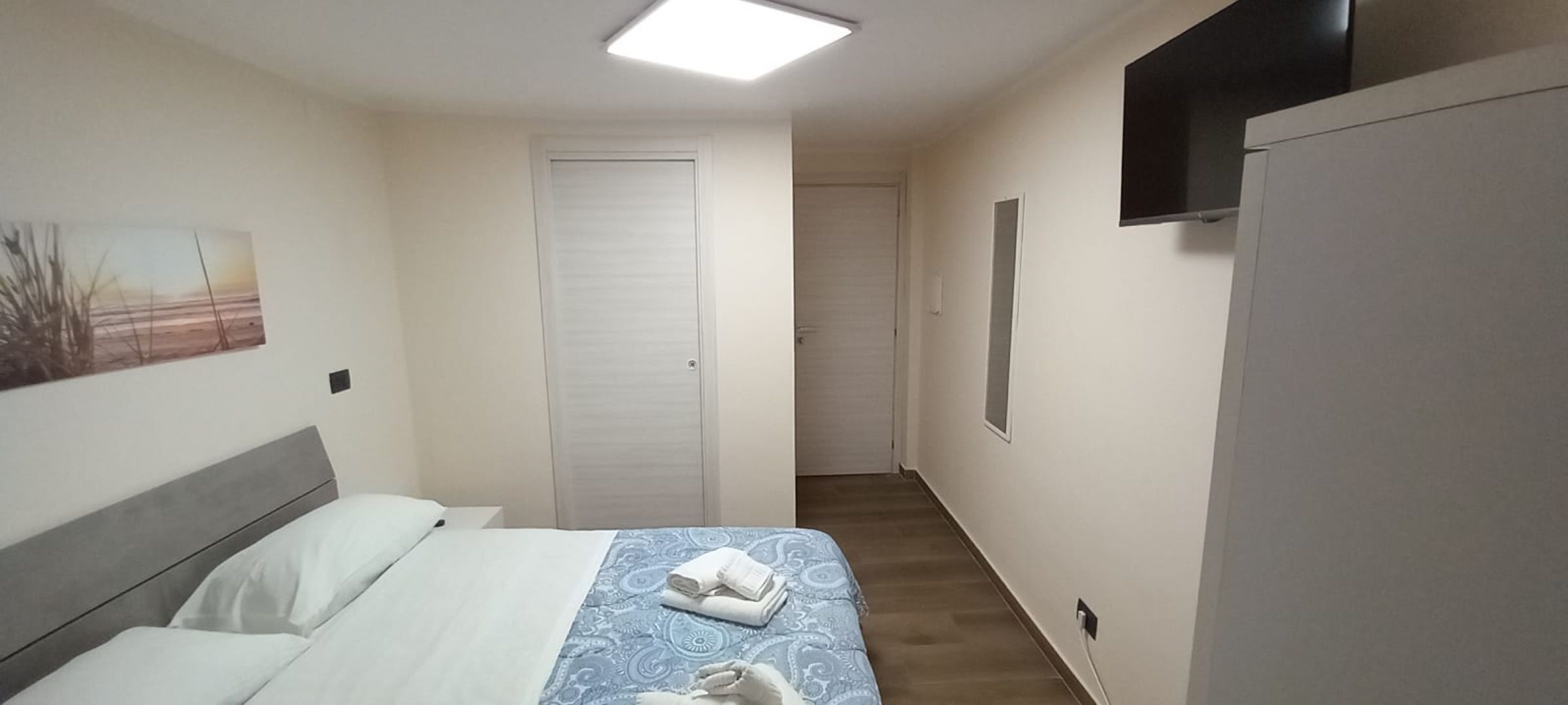 Ein Hotelzimmer mit Bett, einem an der Wand befestigten Fernseher und einer Tür zu einem anderen Raum.