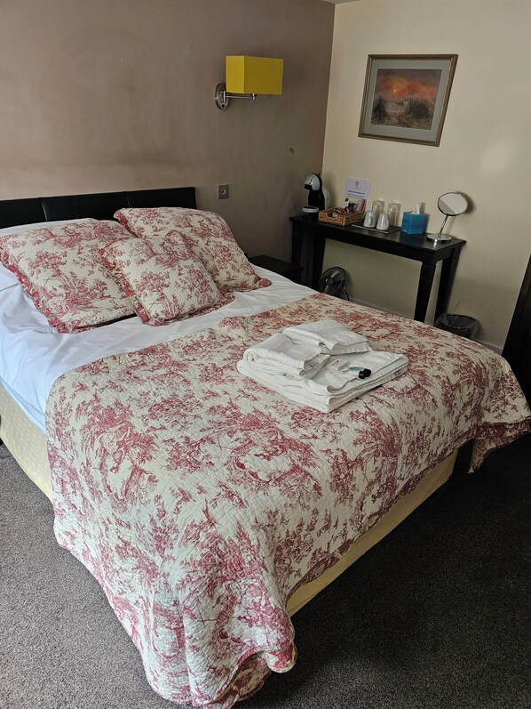 Una cama bien hecha con ropa de cama floral, una mesita de noche con una lámpara y un cuadro en la pared.