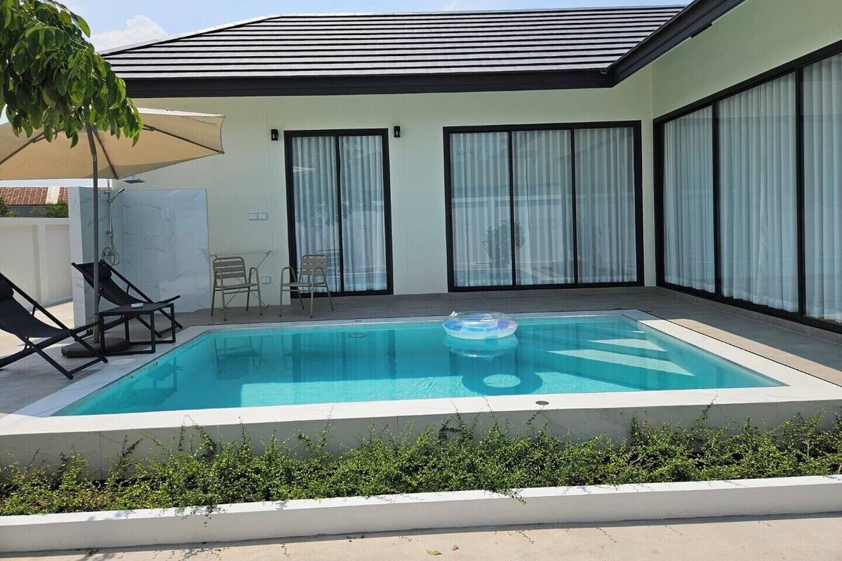 Ein moderner Außenpool mit klarem, blauem Wasser, umgeben von einer niedrigen weißen Mauer, und ein überdachter Sitzbereich mit Stühlen.