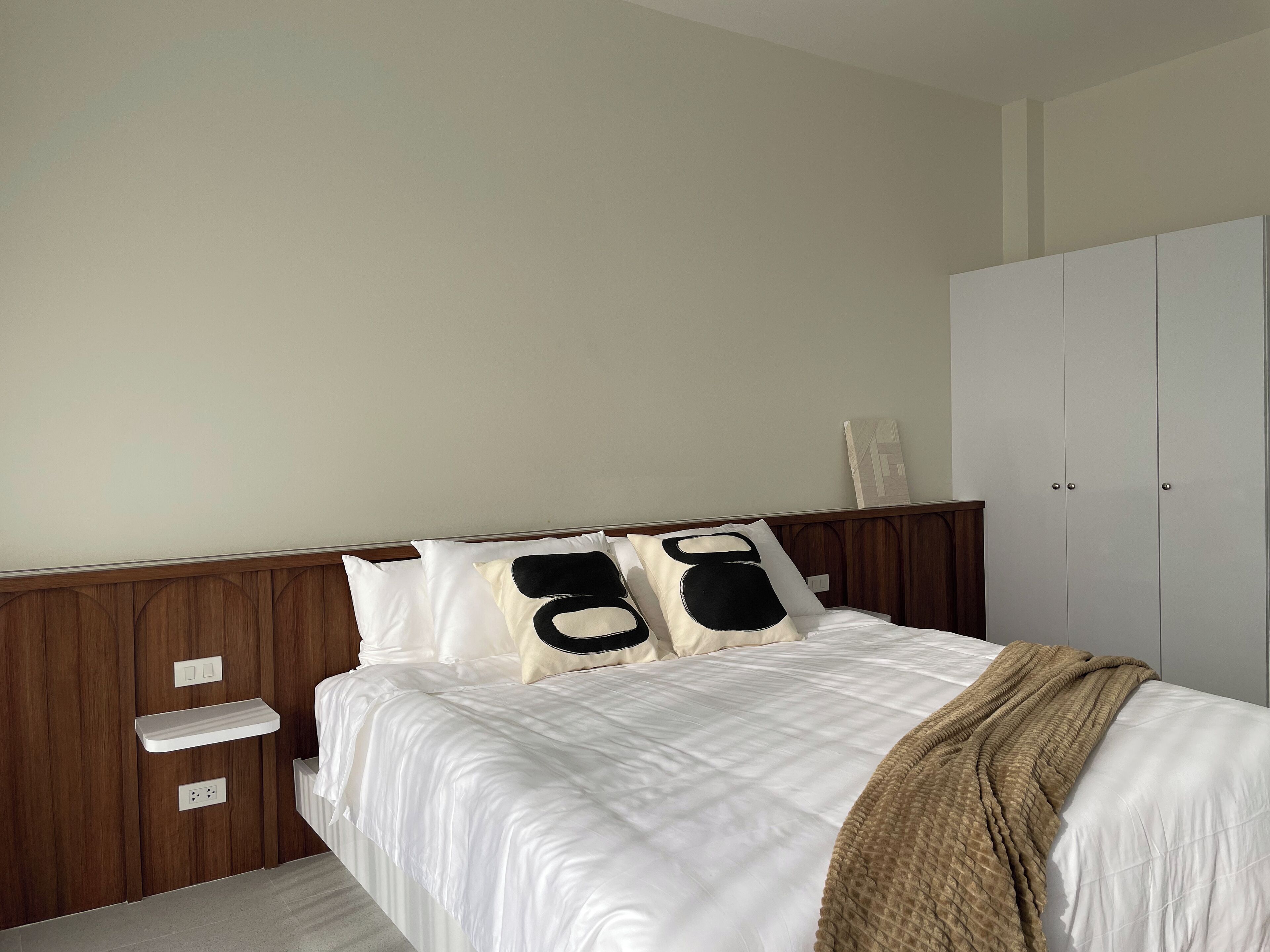 A neatly made bed with white bedding and a brown throw blanket, a wooden headboard, and a white wardrobe with a mirror.