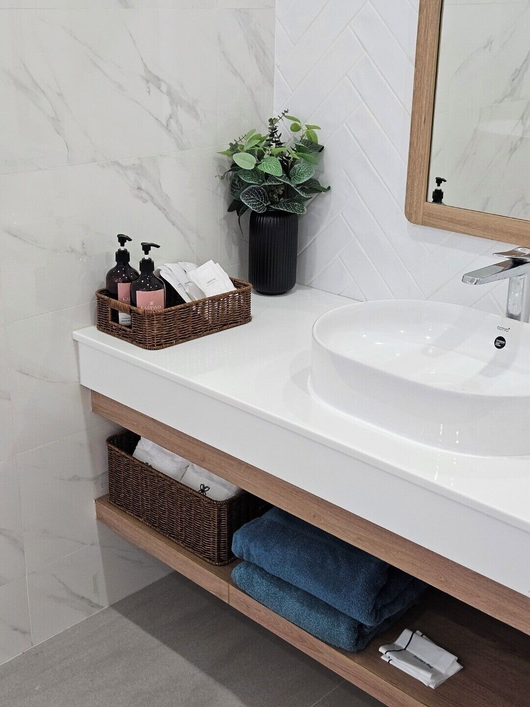 Une salle de bain avec un mur recouvert de marbre blanc, un lavabo blanc, un meuble-lavabo en bois, une plante en pot et un miroir.
