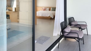Une chambre d’hôtel moderne dotée d’une porte vitrée donnant sur un balcon, d’un lit avec une tête de lit en bois et de deux fauteuils confortables.