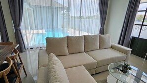 Un salon moderne avec un canapé d’angle beige, une table basse en verre et un coin repas comprenant une table et des chaises en bois.