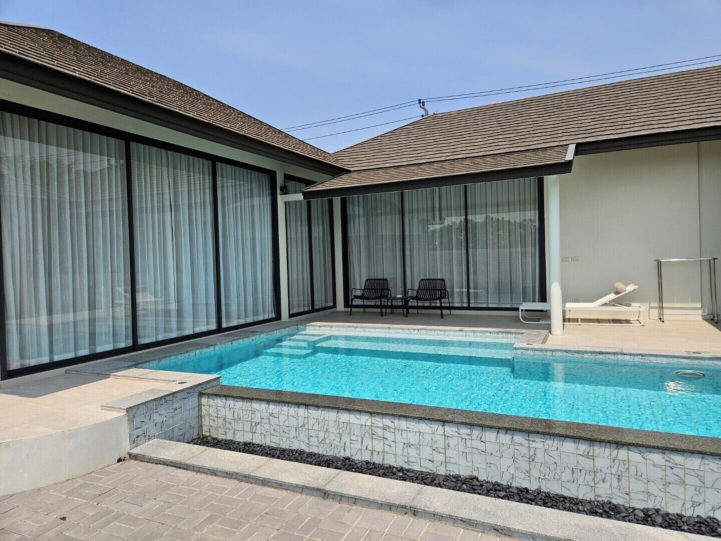 A swimming pool with a tiled edge, two lounge chairs, and a sunbed.