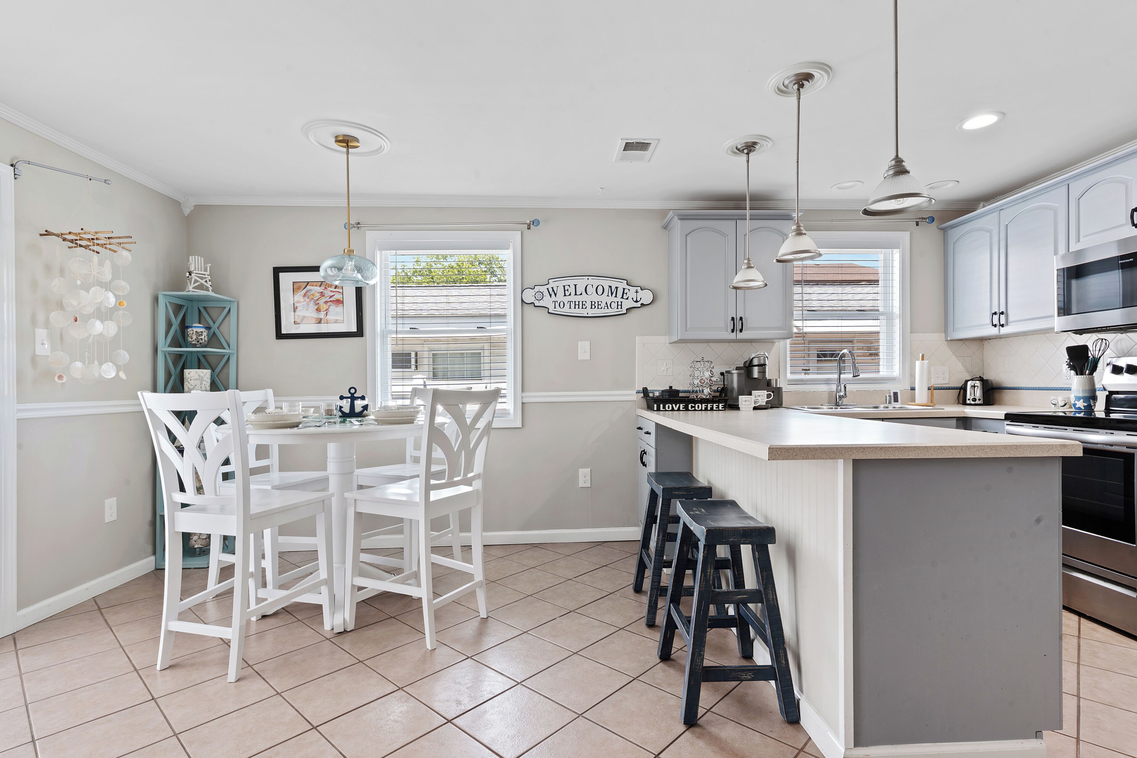 A kitchen with white cabinetry, a central island with stools, pendant lighting, and a 'Welcome to the Beach' sign.