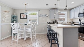 Una cocina con muebles blancos, una isla central con taburetes, iluminación colgante y un letrero que dice 'Welcome to the Beach'.