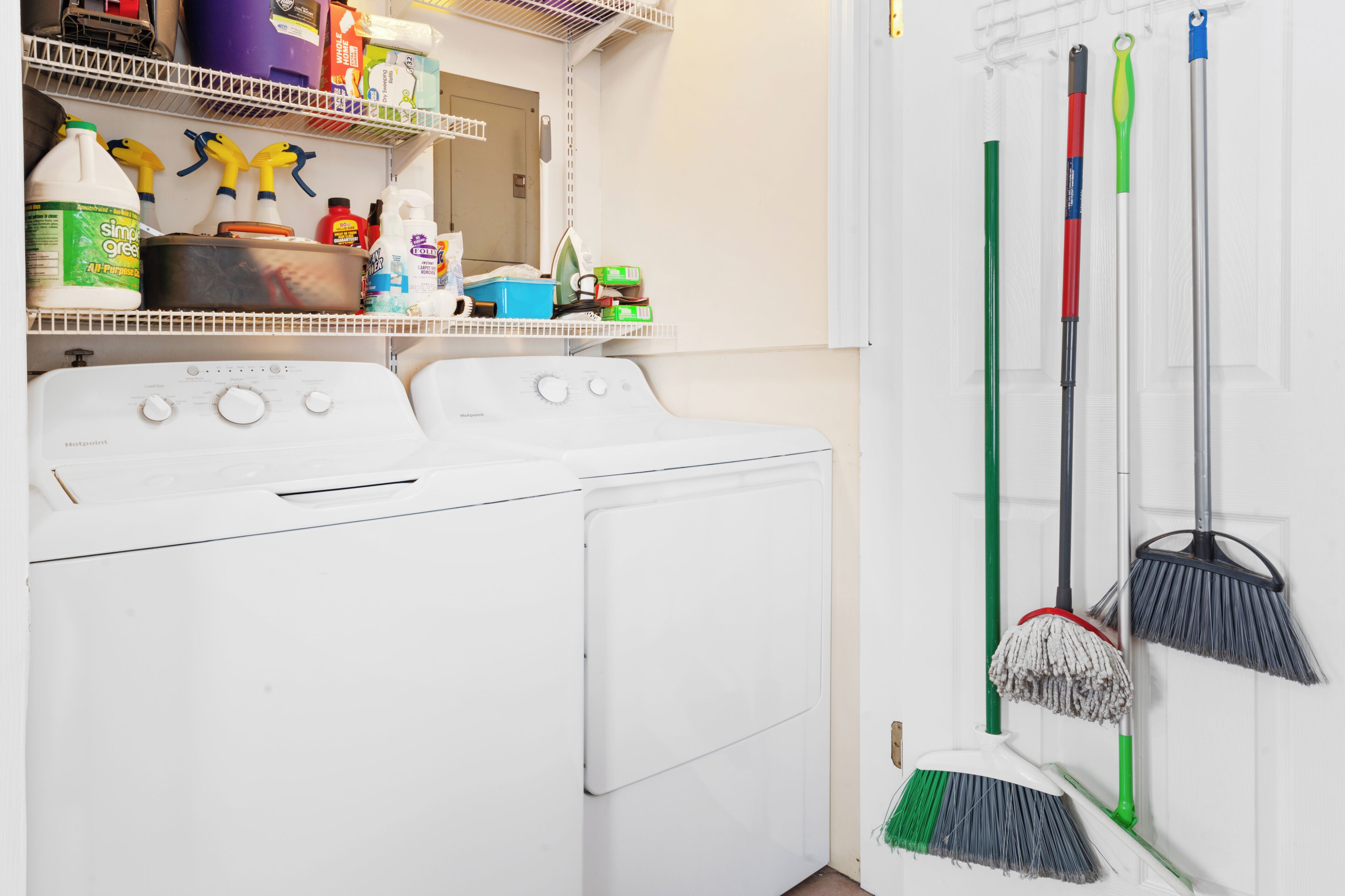 A laundry area with a washing machine, shelves with cleaning supplies, and brooms hanging on the wall.
