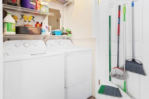A laundry area with a washing machine, shelves with cleaning supplies, and brooms hanging on the wall.