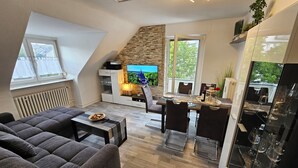 A modern living room with a sofa, dining table, and TV. There is a fireplace, a window with curtains, and a plant on the wall. - Bright and modern apartment in the attic very close to the centre and university (Kassel)