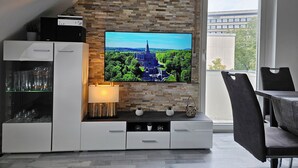 A modern living room with a stone accent wall, a flat-screen TV, a dining area with a table and chairs, and a glass cabinet with decorative items. - Bright and modern apartment in the attic very close to the centre and university (Kassel)