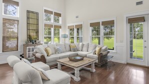 A spacious living room with a large sectional sofa, a coffee table, and a view of a green lawn through large windows. - Two Golf Carts ~ Golf Course Views ~ Pet Friendly (Miramar Beach)