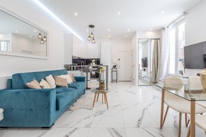 A modern living room with a blue sofa, a glass coffee table, and a dining area with a glass table and chairs. - Renovated Apt Min. frm Beach/Palais - 30m2 Terrace (Cannes)