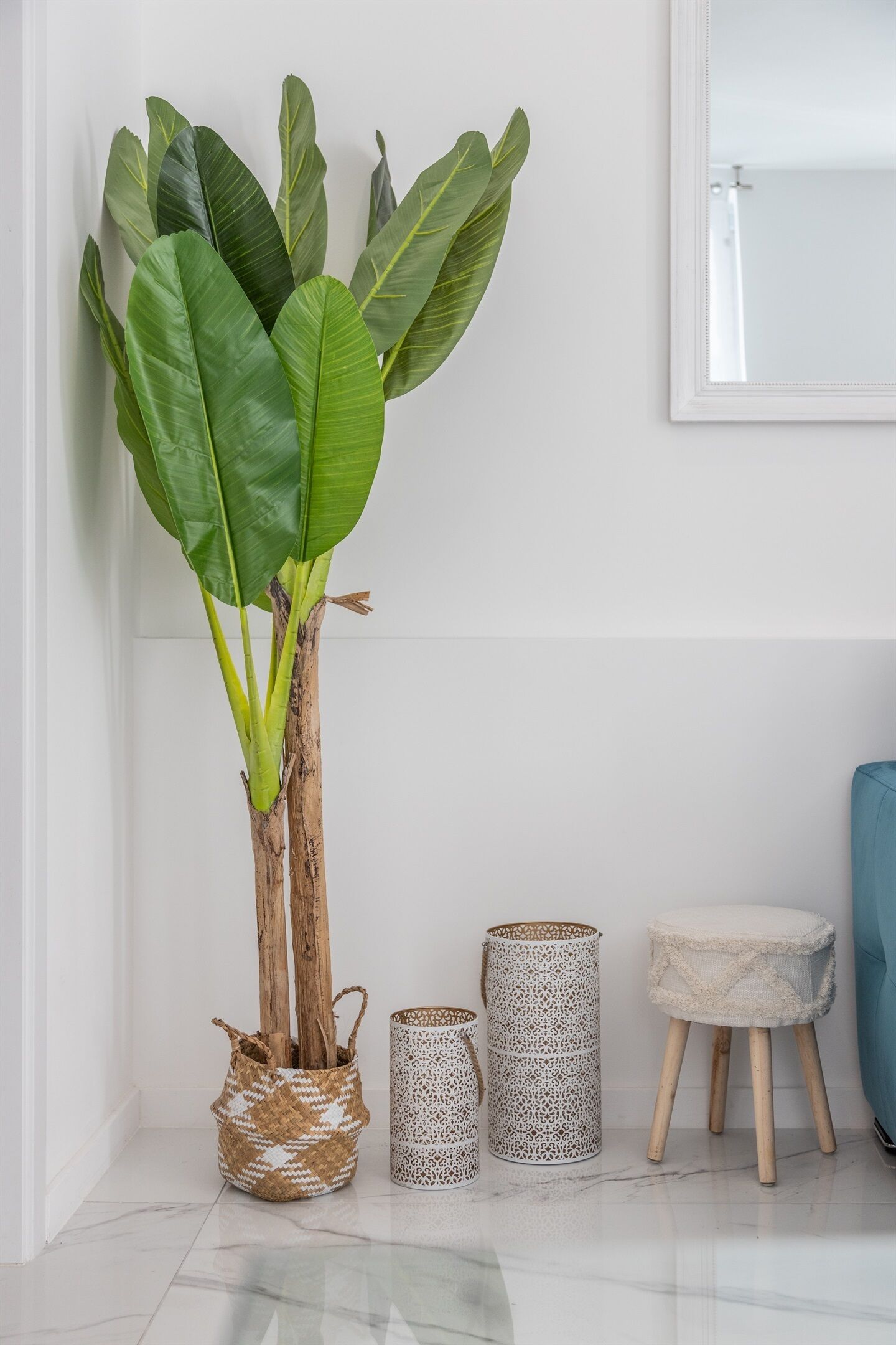 Una planta en maceta de hojas grandes, dos cestas tejidas y un taburete pequeño contra una pared blanca.