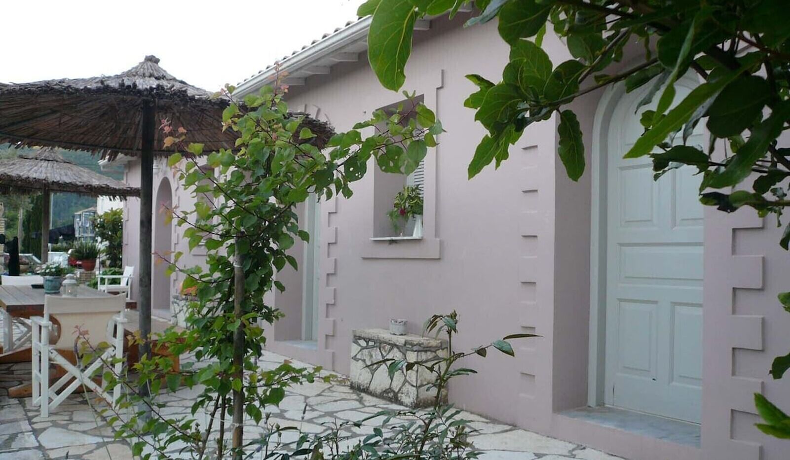Un edificio rosa con un patio de piedra, una estructura de techo de paja y una puerta blanca.