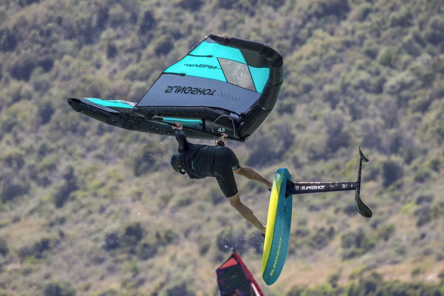 Uma pessoa praticando kitesurf com uma prancha com a inscrição "SLINGSHOT" e uma pipa com os logotipos "TOMAS" e "TOMASHOL".
