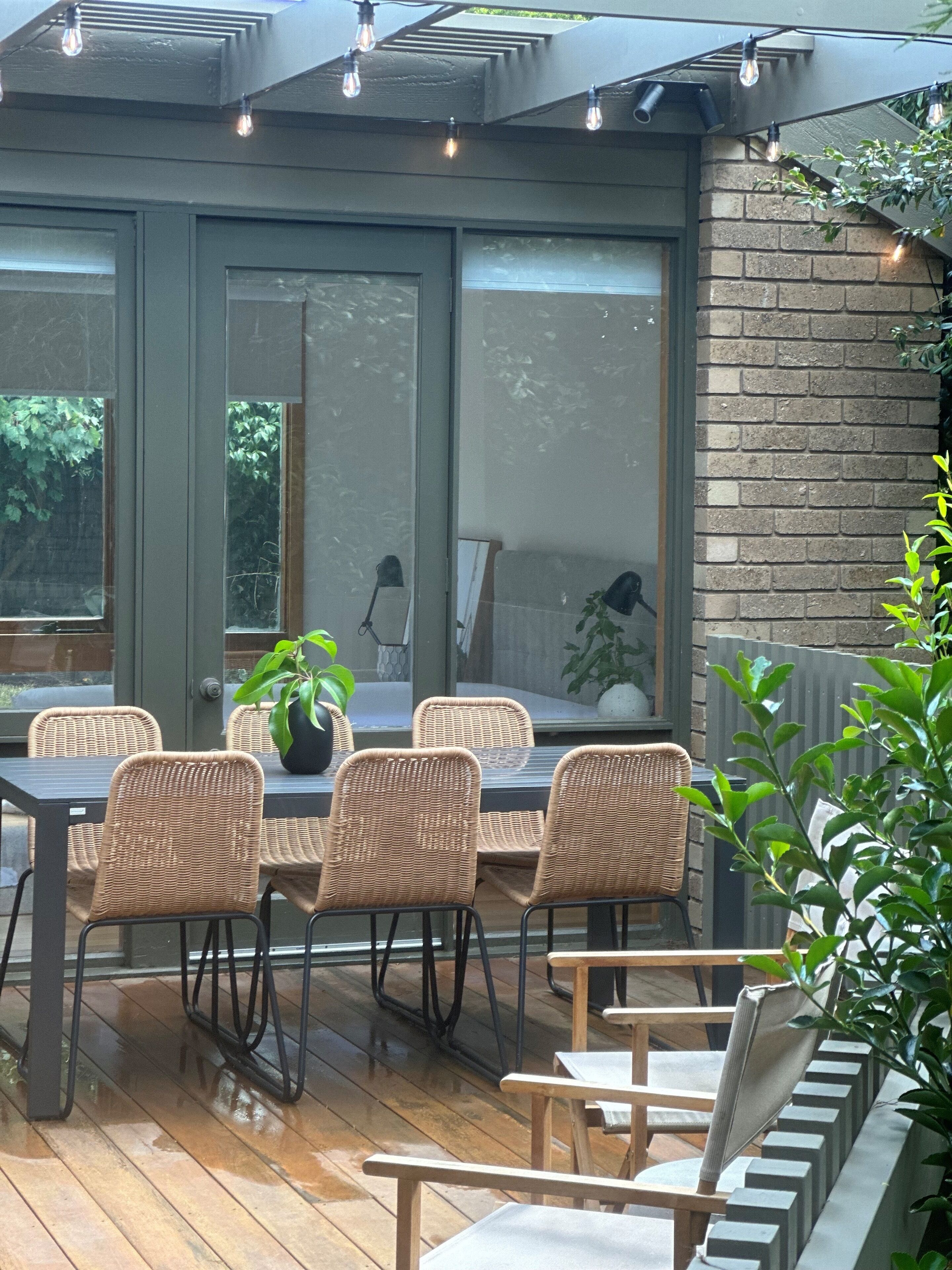 Un patio con sillas de mimbre y una mesa, ventanas amplias y una pared de ladrillo.