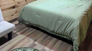 Una habitación con paneles de madera, una cama con colcha verde, un almohádega azul, una mesita de noche con una lámpara y una ventana con cortinas.