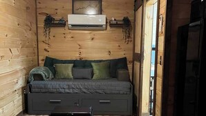 Acogedor cuarto estilo cabaña con interior de madera, un sofá cama con almohadas, una mesita y un aire acondicionado instalado en la pared.