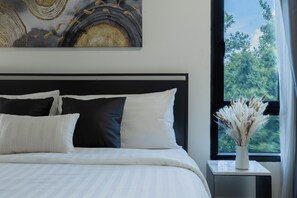 A neatly made bed with white and black bedding, a small bedside table with a vase of dried flowers, and a large window with a view of greenery. - NORN Riverside Bangkok Hotel (Bangkok)