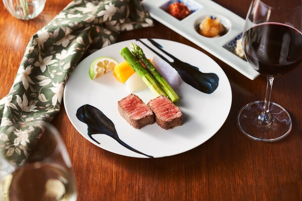 Un plato de carne, espárragos, limón y salsa, acompañado de una copa de vino tinto y un mantel floral.