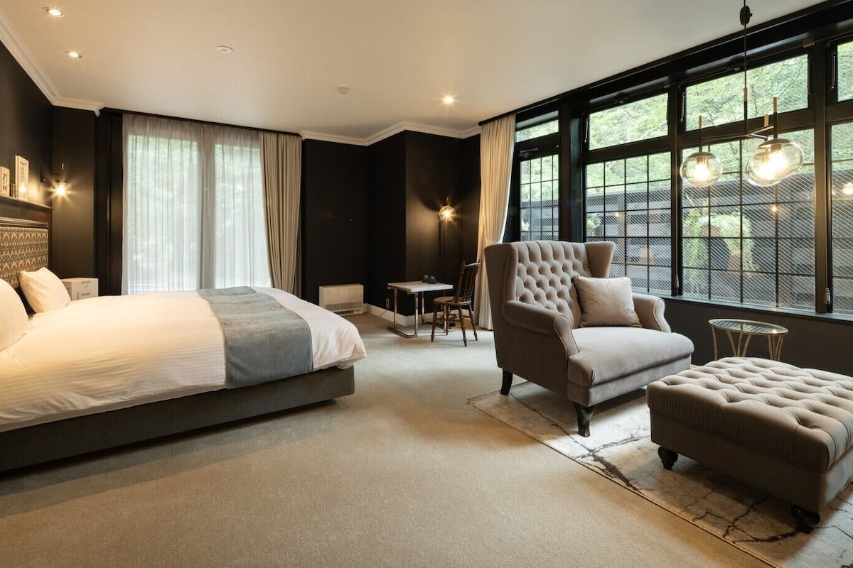 A modern bedroom with a large bed, a sofa, an ottoman, and a dining area.