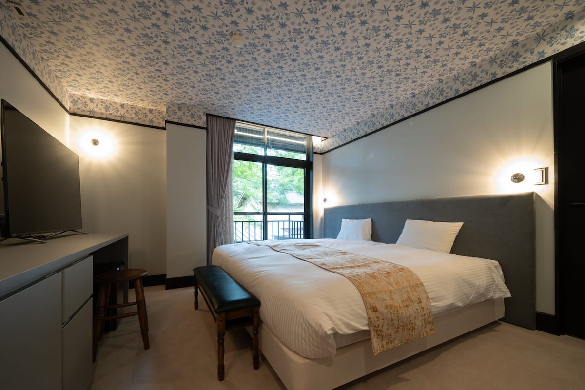 A bedroom with a large bed, a desk, and a chair. There is a window with curtains and a view of greenery outside.