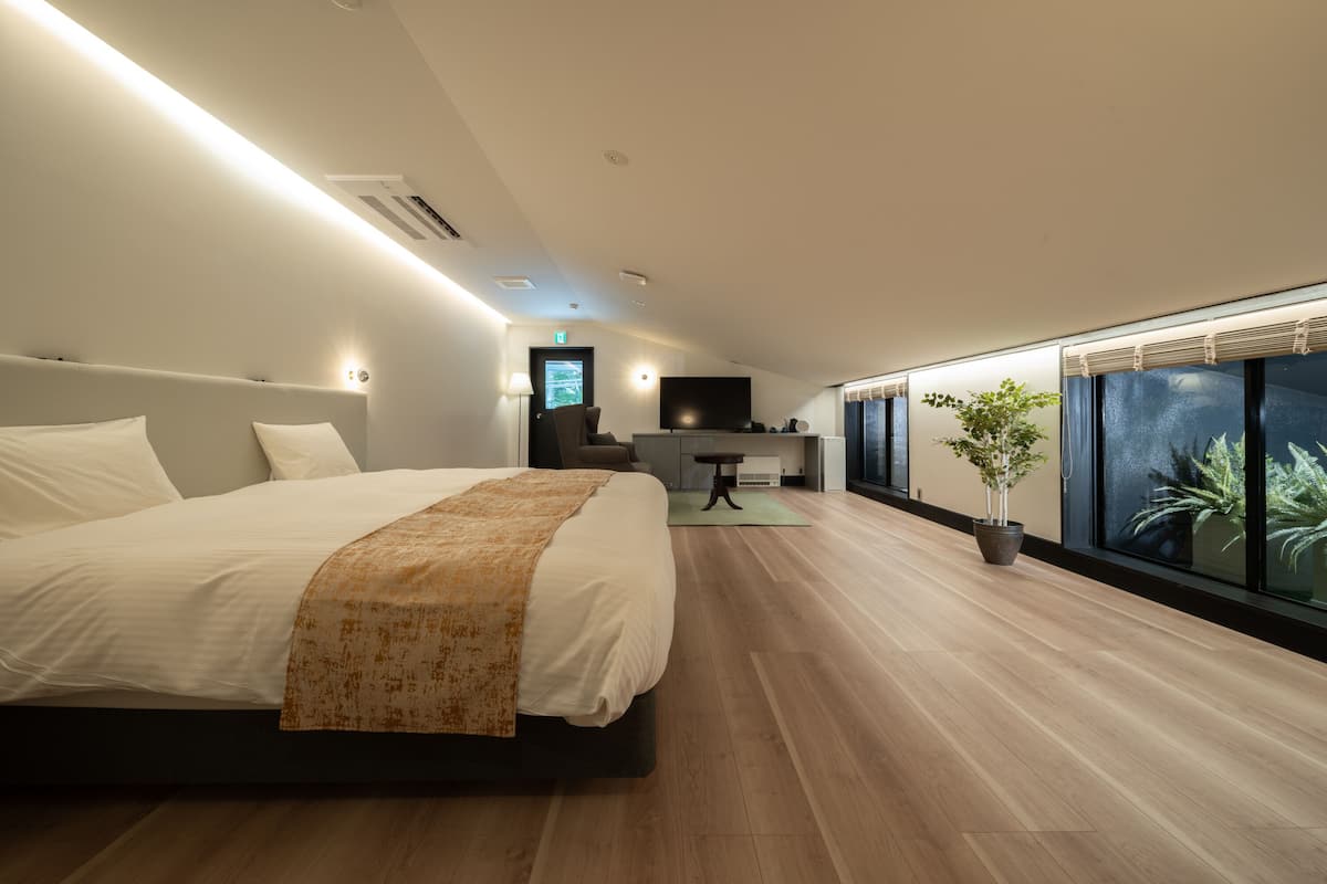A modern hotel room with a large bed, a small seating area, a TV, and a large window with a view.