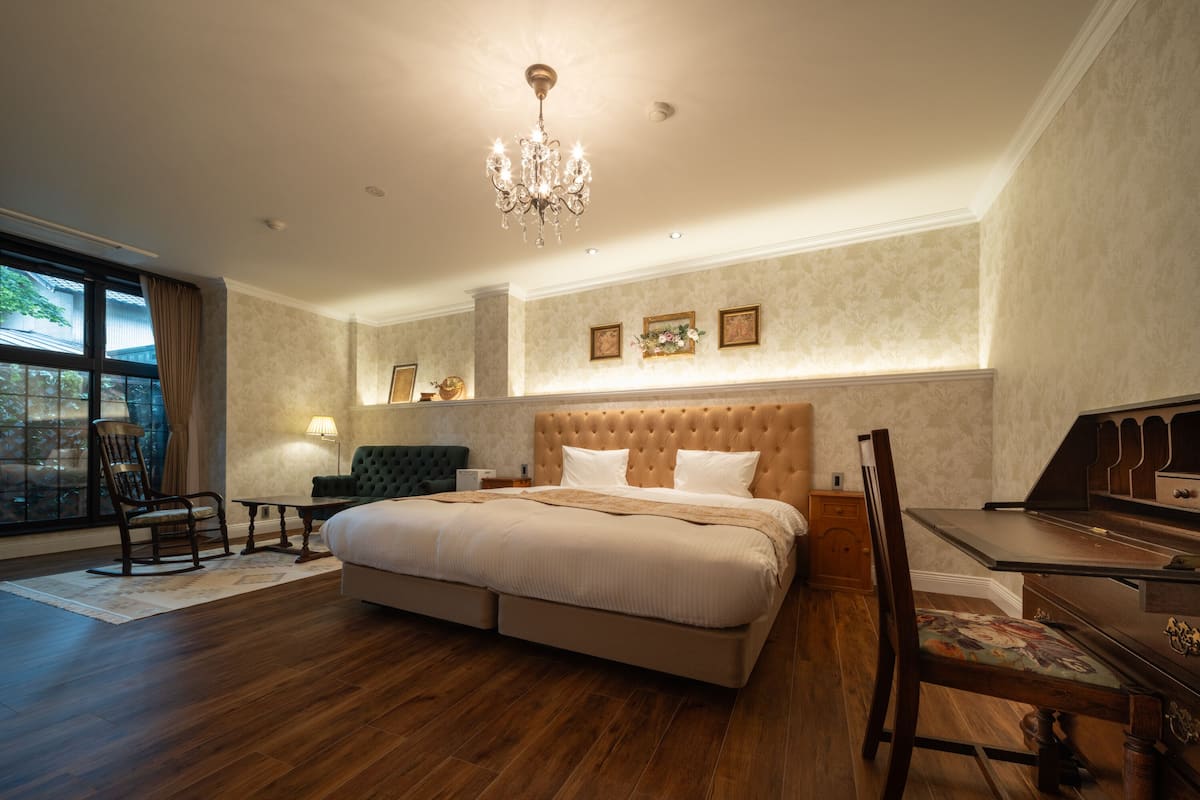 A spacious bedroom with a large bed, a desk, and a chair. There is a chandelier hanging from the ceiling and a window with curtains.