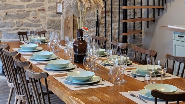 A dining area with a long wooden table set for a meal, surrounded by chairs. A stone wall and a staircase with black railings are visible in the background.