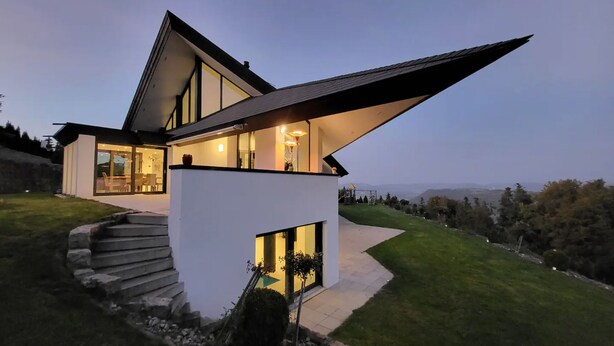 A modern house with a unique sloped roof, large windows, and a staircase leading to a covered porch. - Hilltop Luxury Villa with Minigolf, Indoor Pool, Wellness & Spa (Oberbütschel)