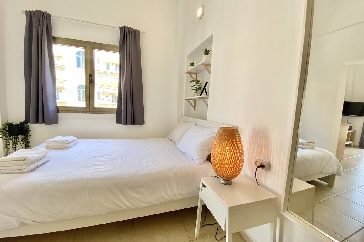 Un lit bien fait, avec du linge de lit blanc, et une petite table de chevet sur laquelle se trouve une lampe.