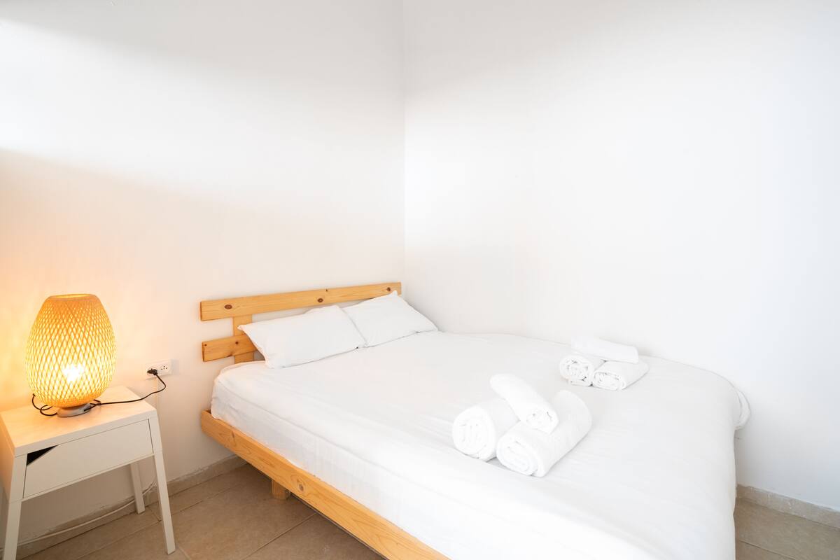 Un lit bien fait, avec du linge de lit blanc et des serviettes pliées, une lampe de chevet et une tête de lit en bois.