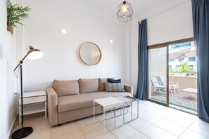 A modern living room with a sofa, a coffee table, a floor lamp, a round mirror, and a sliding glass door leading to an outdoor area with chairs.