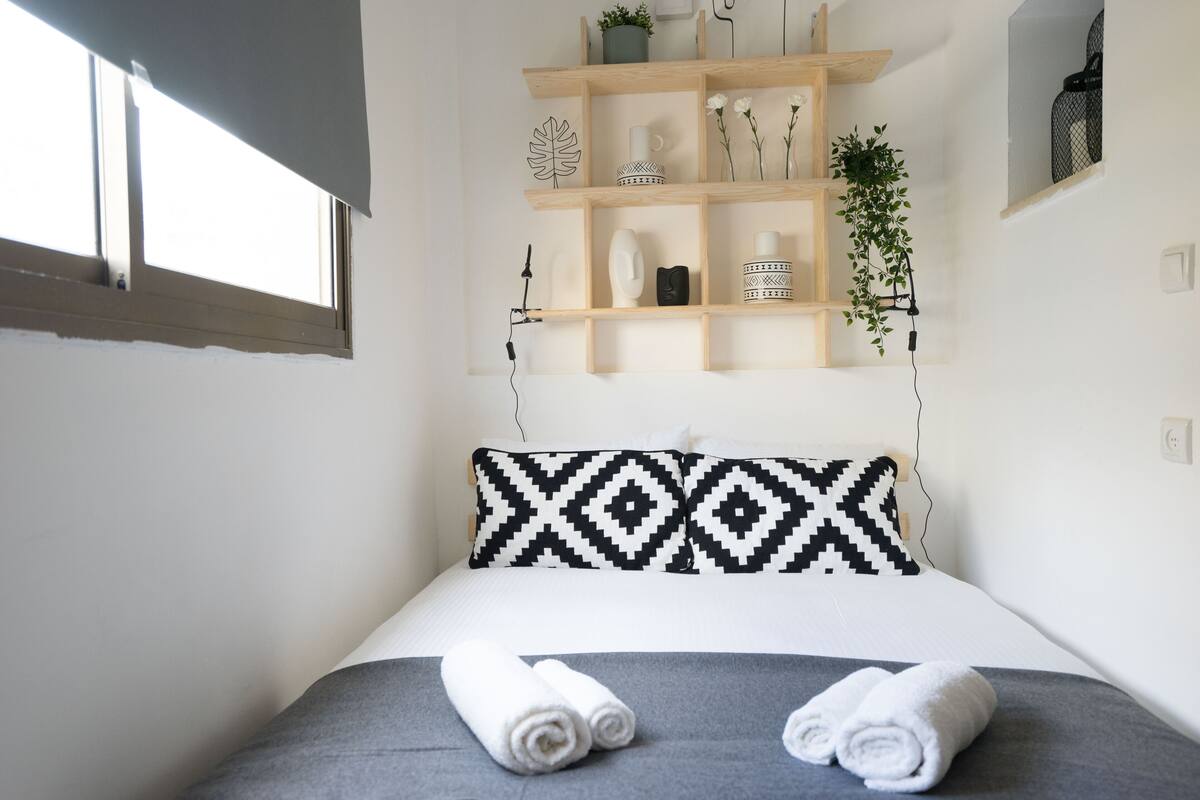Un lit bien fait, avec deux serviettes enroulées, une étagère en bois ornée d’objets décoratifs et une fenêtre avec des stores.