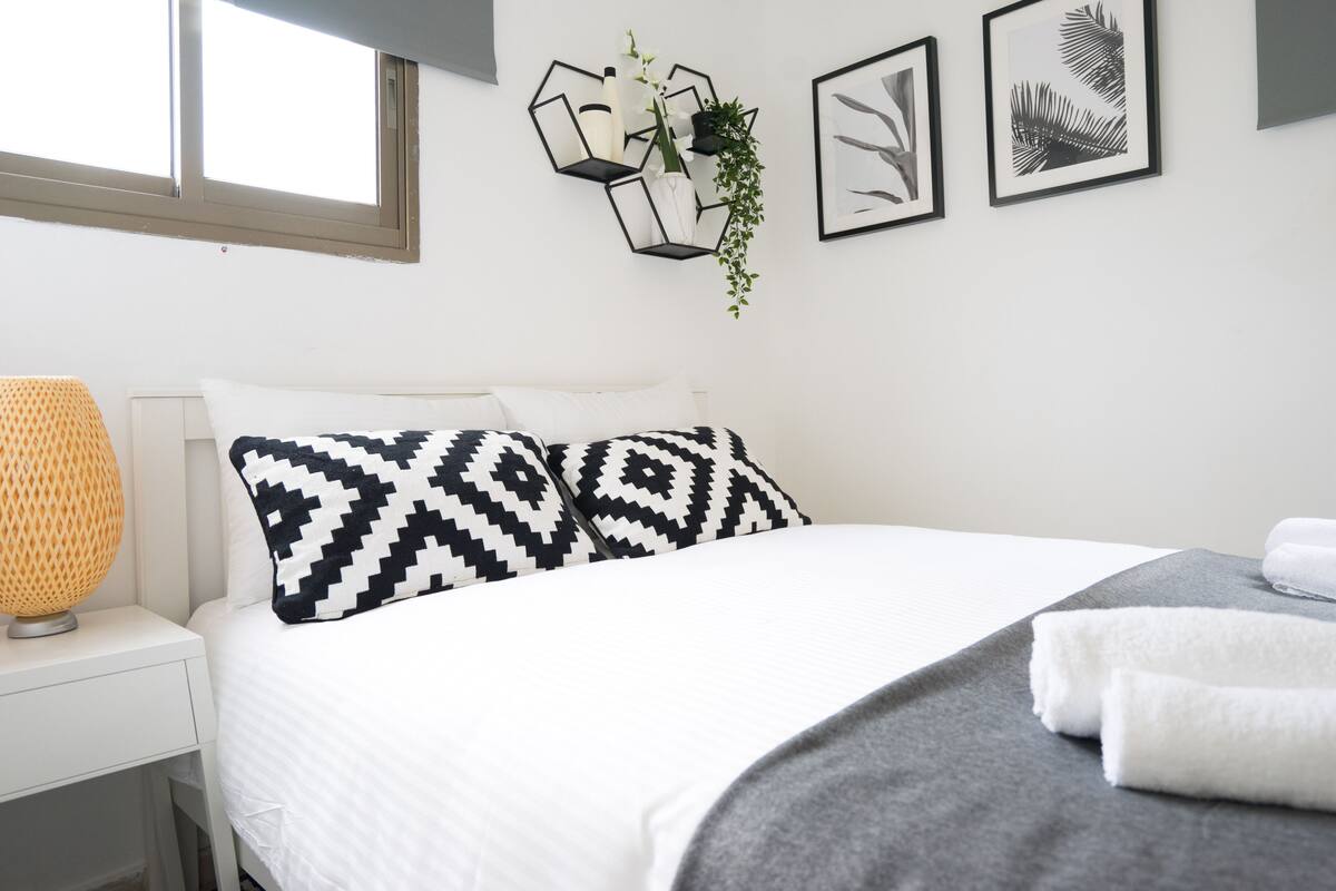 Une chambre à coucher bien rangée, avec un lit, du linge de lit blanc, une lampe de chevet et des éléments de décoration fixés au mur, notamment des œuvres d’art encadrées et une étagère géométrique agrémentée de plantes.
