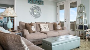 A living room with a beige sofa, a light blue ottoman, a lamp, a decorative mirror, and a 'BEACH' sign on the wall.