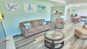 A spacious living room with a beige sofa, a round coffee table, and a dining area with a whiteboard. - Casa Rosita 3 Bedroom Home by RedAwning (South Padre Island)
