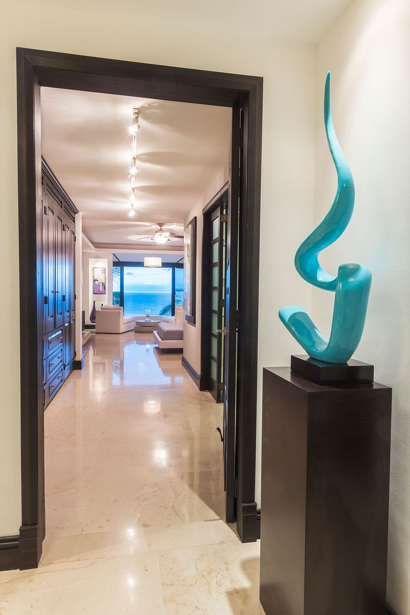 A hallway with a modern design, a sculpture on a pedestal, and a view to a living area with a large window.