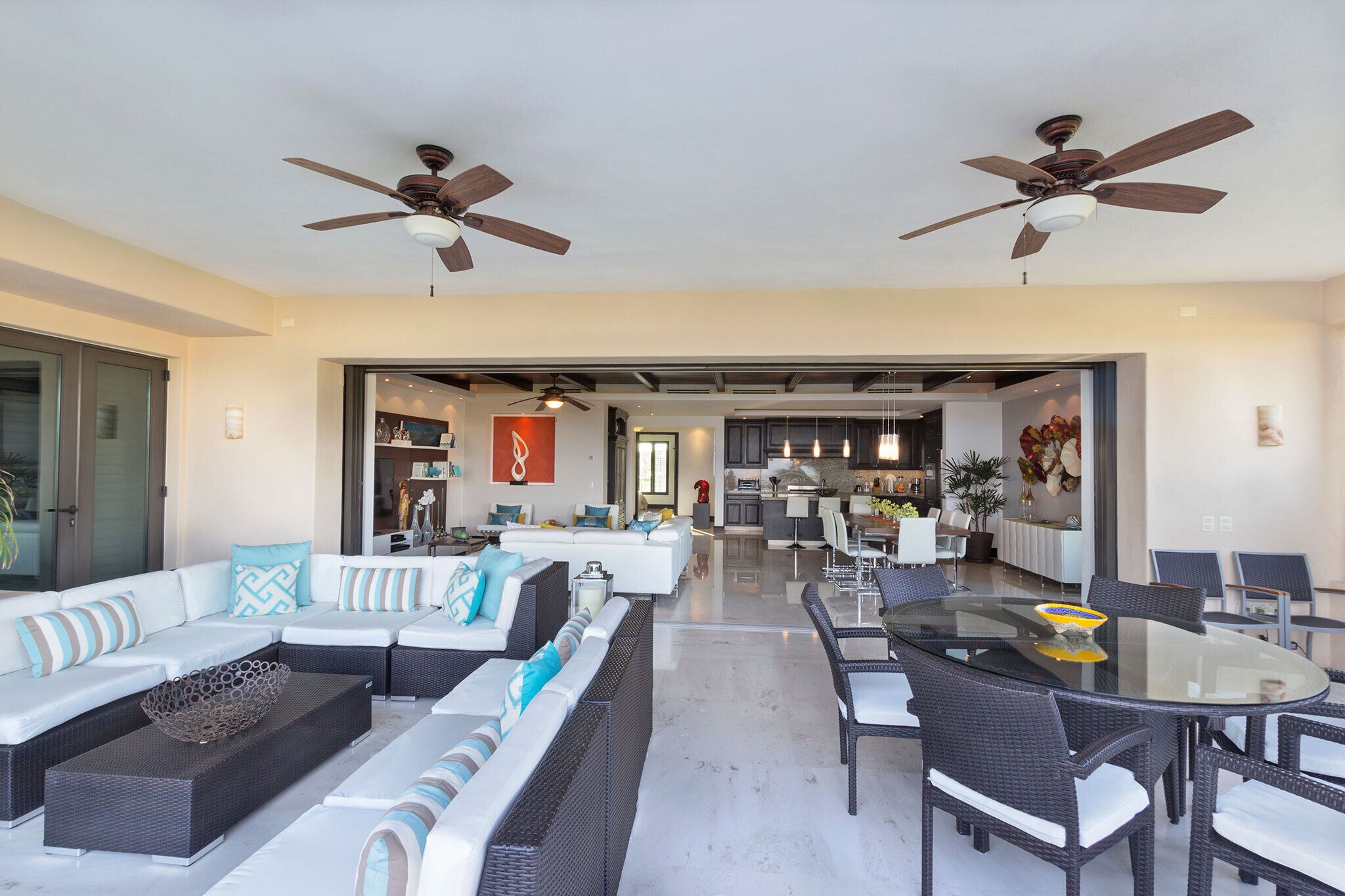 A spacious living area with a glass-top dining table, dark wicker furniture, and a modern kitchen in the background.