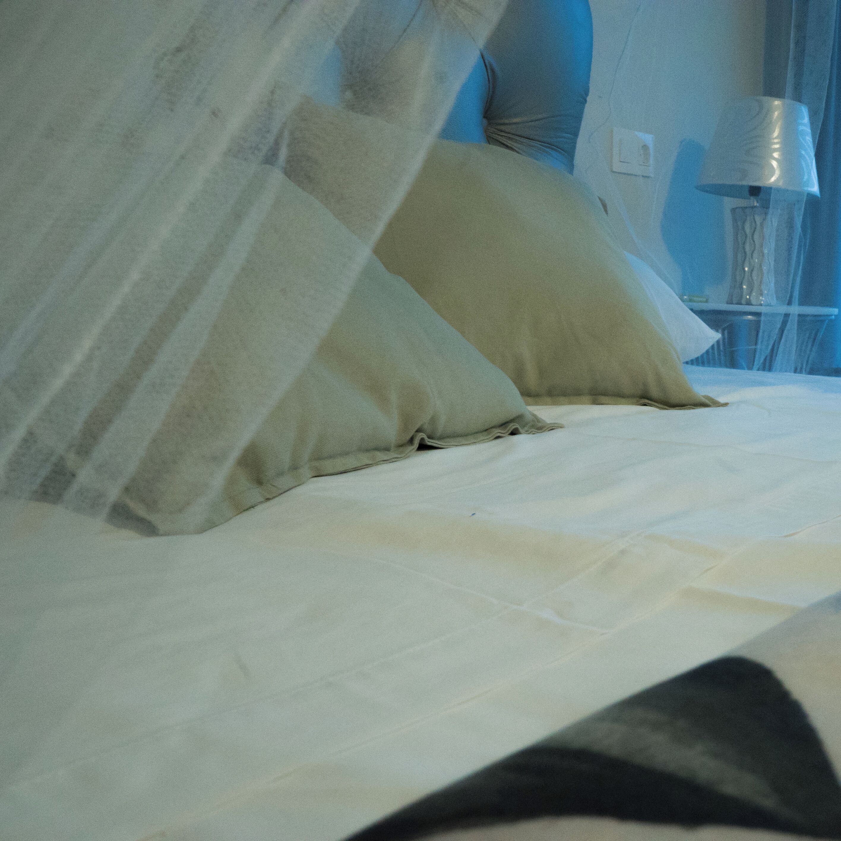 A bed with white linens and two pillows, a bedside table with a lamp, and a window with sheer curtains.