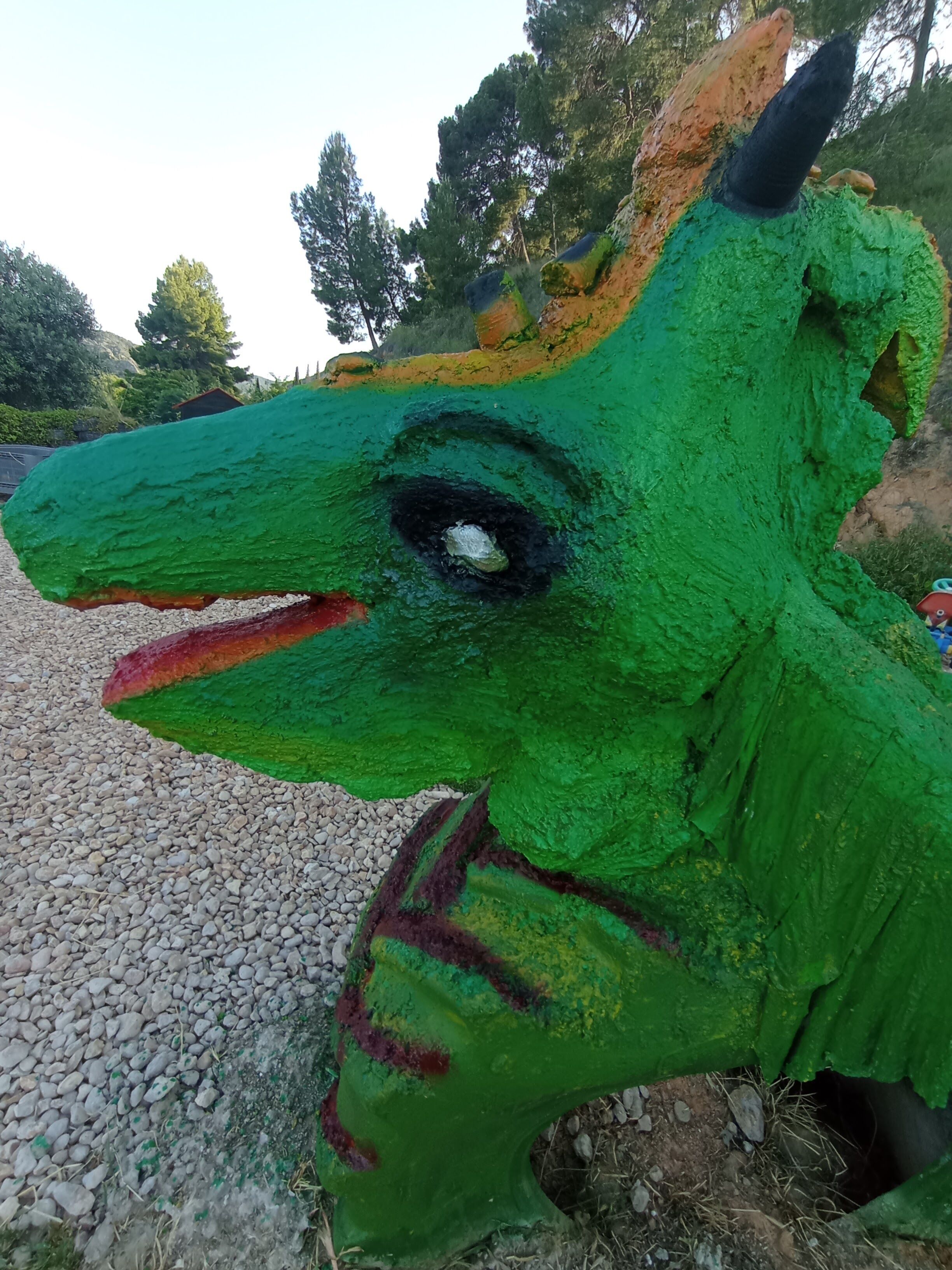 Una escultura de dinosaurio verde con superficie texturizada, ubicada en un camino de grava con árboles al fondo.