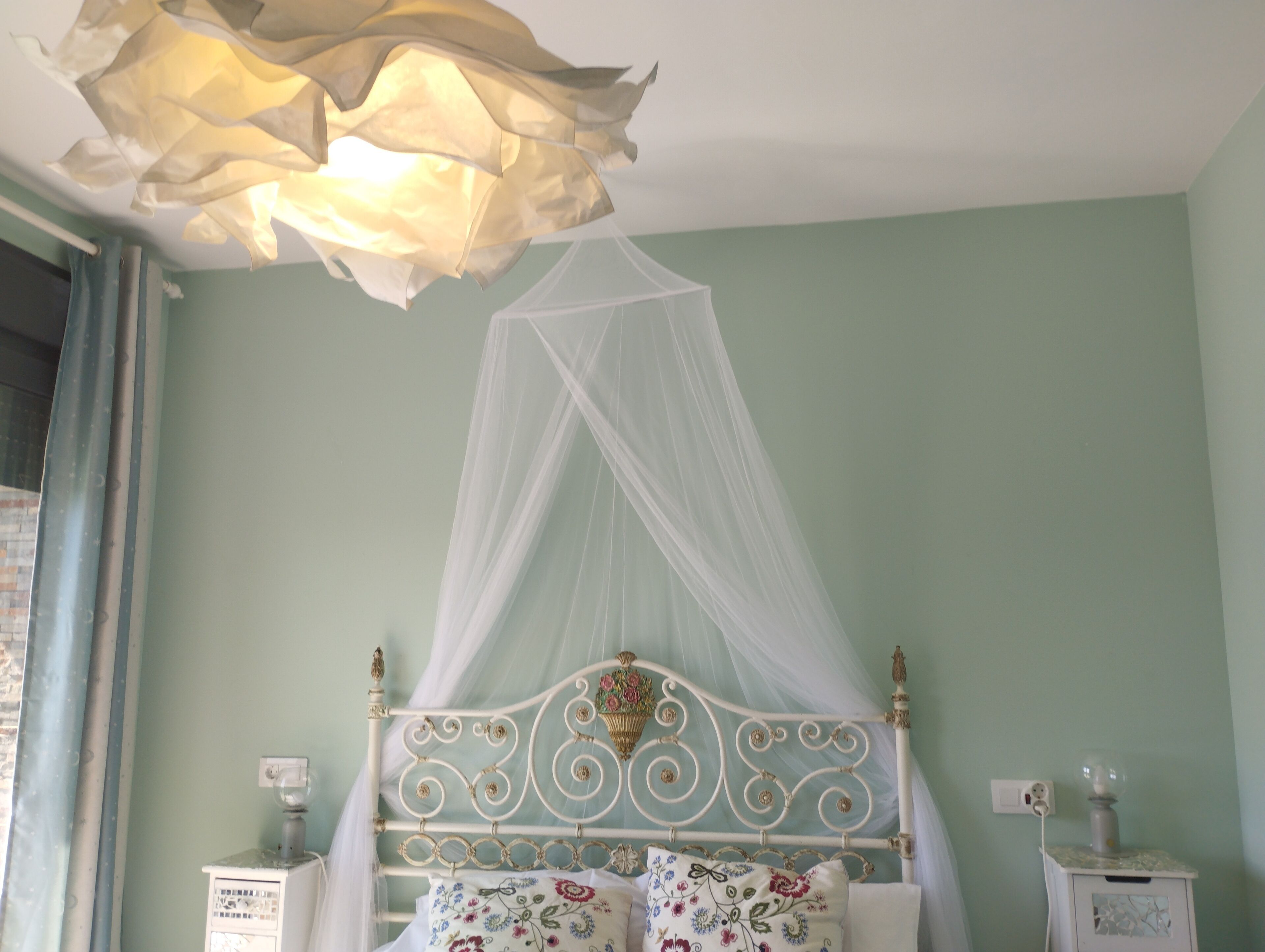 A bedroom with a bed under a white canopy, a nightstand with a lamp, and a wall socket.