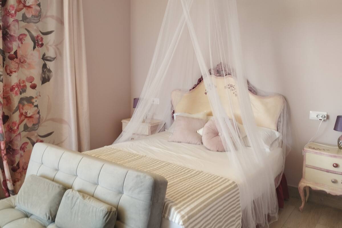A bedroom with a canopy bed, a bedside table, a lamp, and a floral curtain.